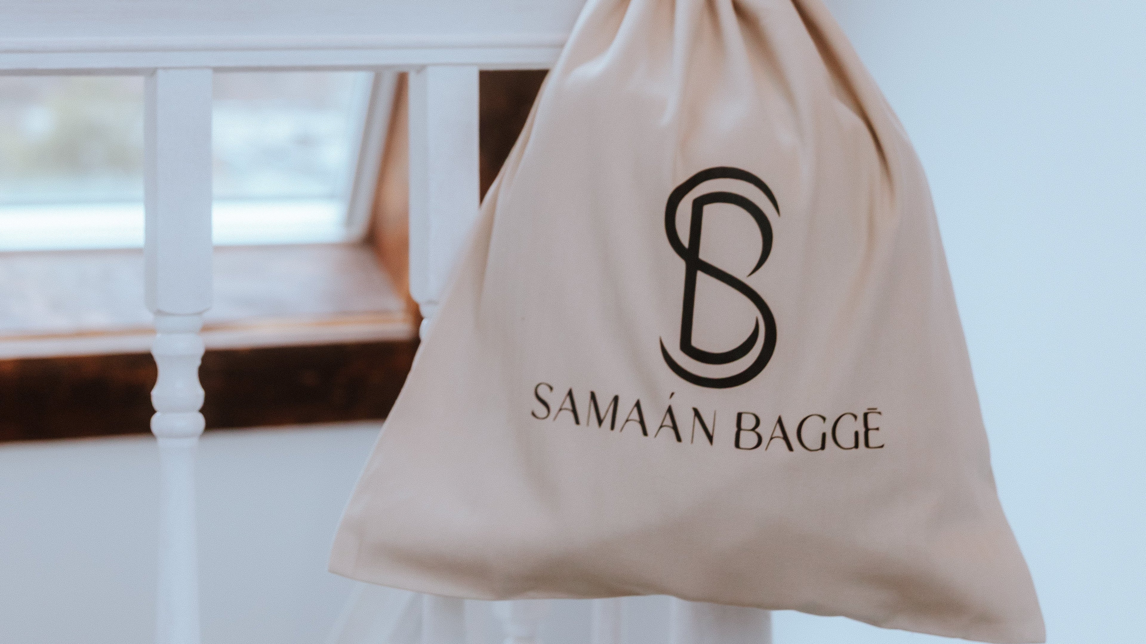 Beige drawstring bag with 'Samaan Bagge' logo on a white surface with a window in the background.