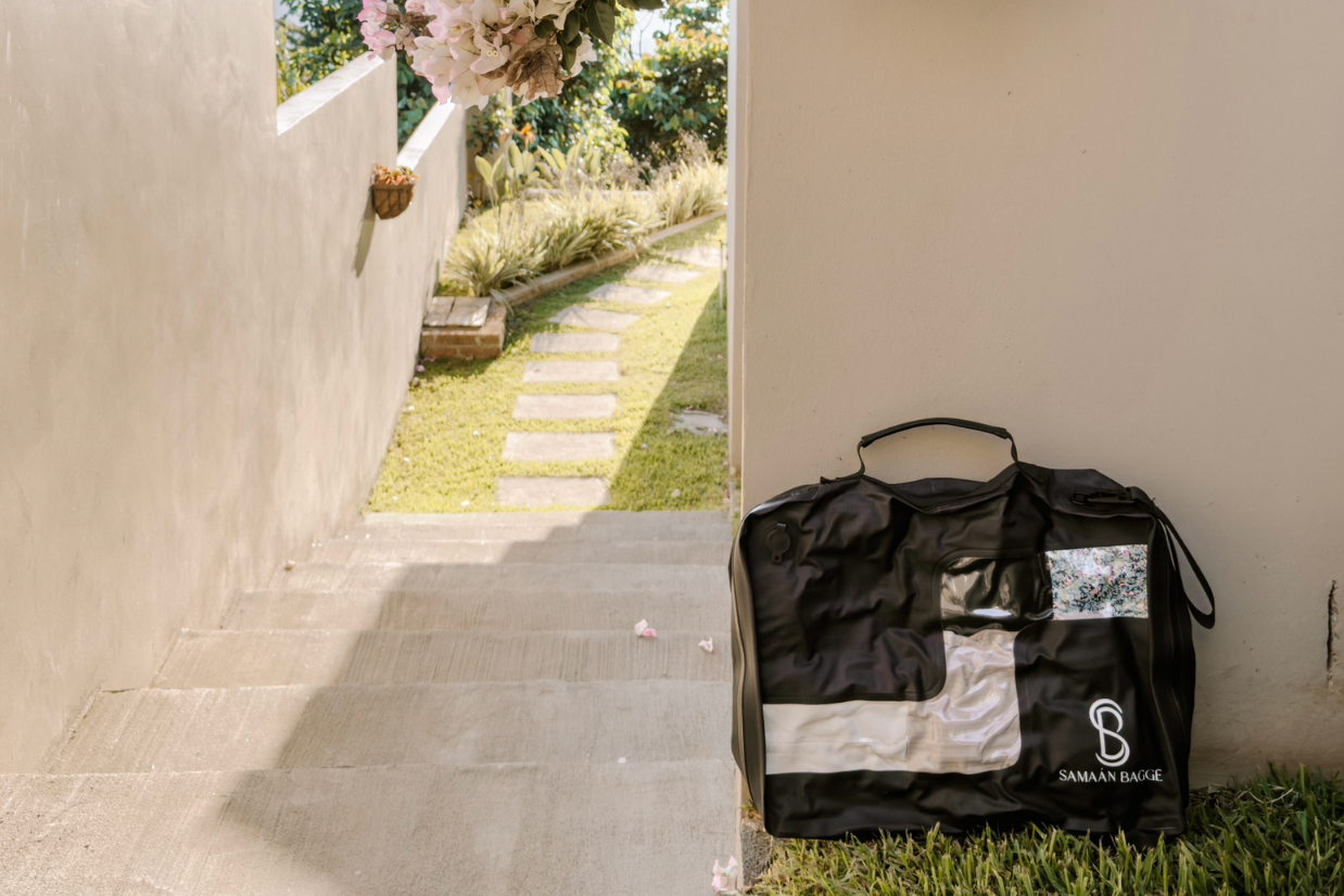 Black packing compression bag with Samaan brand logo on a grassy area near a stone wall and flowers.